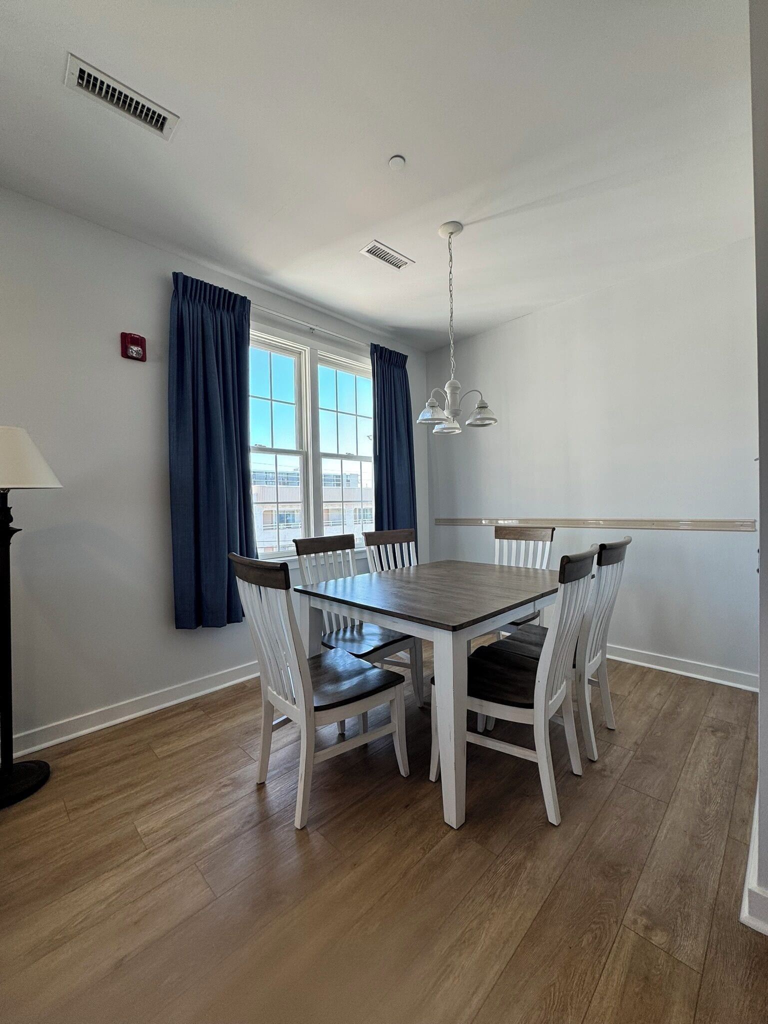 A dining area with a wooden table and white chairs, a window with curtains, and a ceiling fan.
