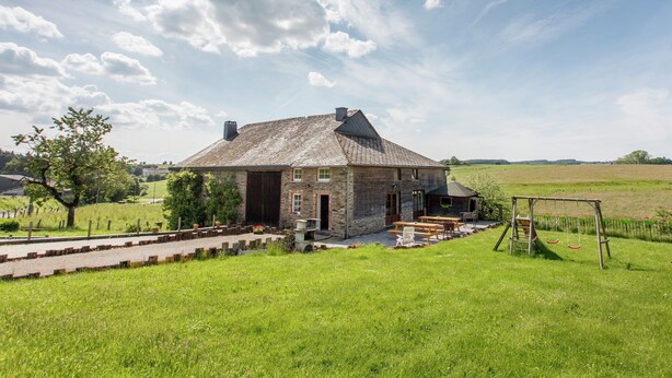 A stone farmhouse with a large porch, surrounded by a grassy field and a clear sky.