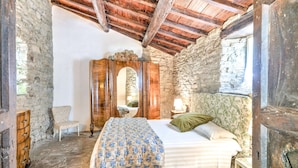 Une chambre à coucher rustique avec un mur en pierre, des poutres apparentes en bois au plafond, un lit recouvert d’une courtepointe à motifs floraux, une armoire en bois et une petite table de chevet avec une lampe.