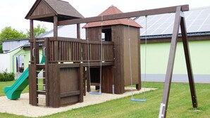 A wooden play structure with a slide and swings, set on a grassy area with a building in the background.