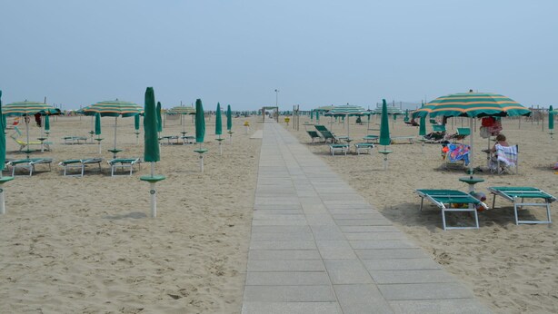 A beach with rows of green and white striped umbrellas, sun loungers, and a paved walkway. - Belvilla by OYO Lido Estensi Trilo Doria (Comacchio)