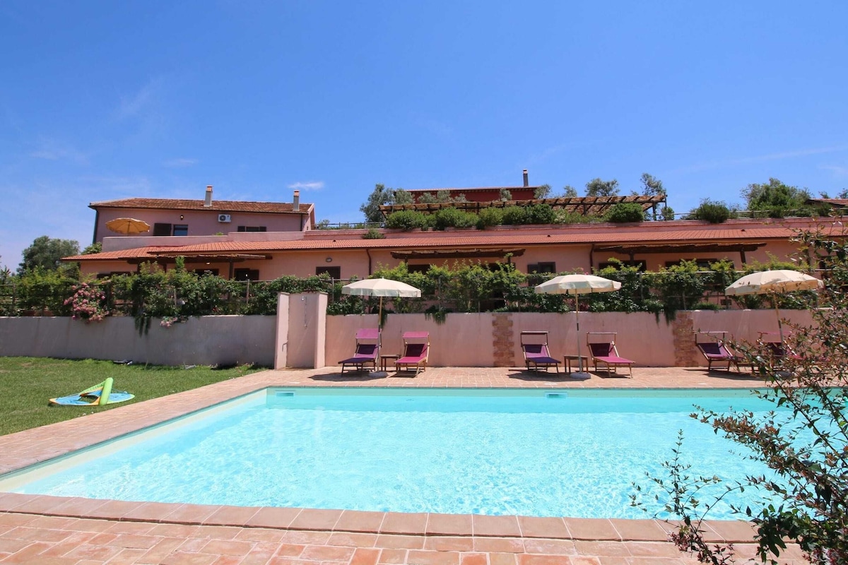 A swimming pool with lounge chairs and umbrellas, surrounded by a landscaped garden and a large building with a red-tiled roof.