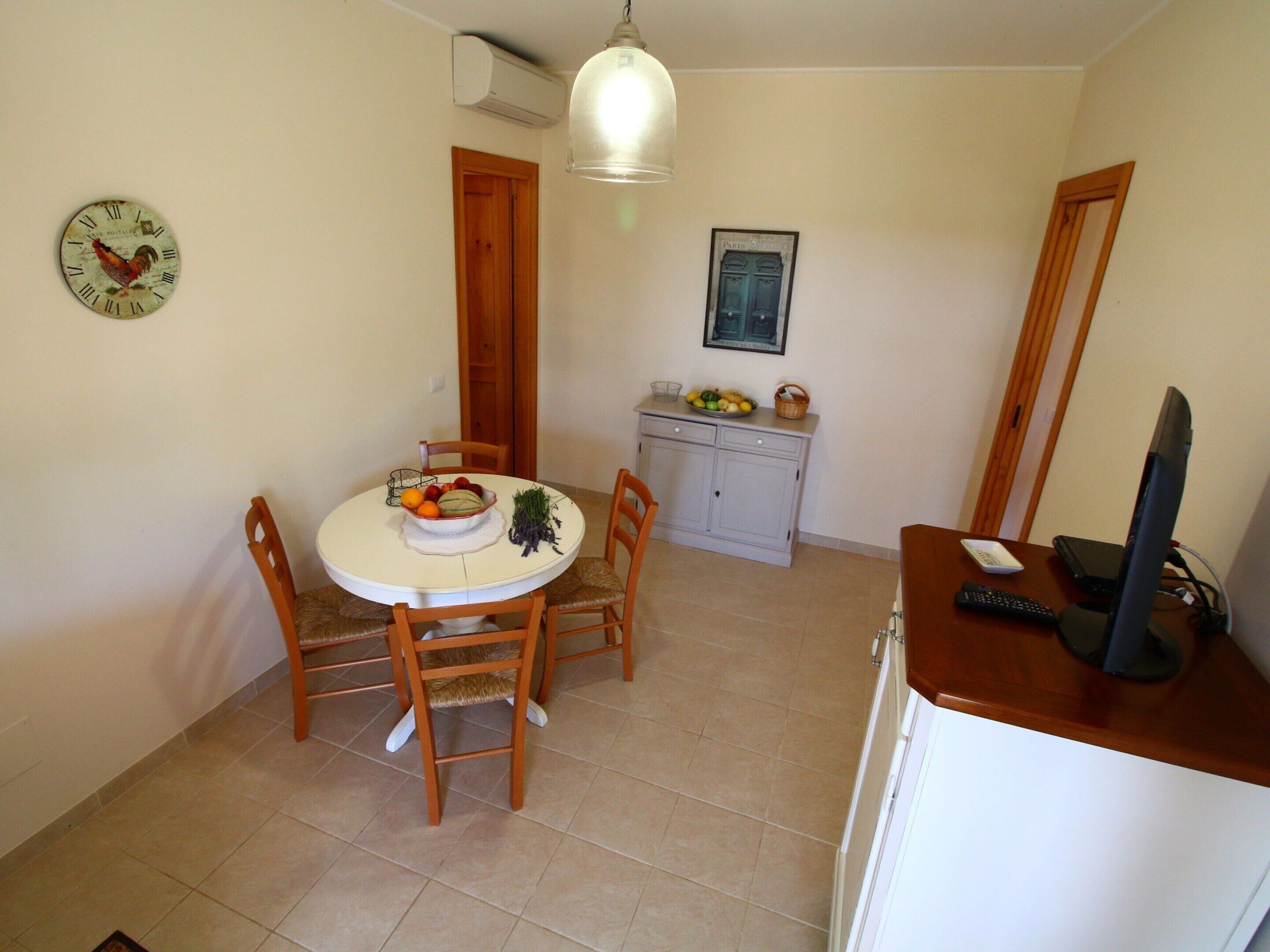 A compact dining area with a round table, wooden chairs, a small cabinet with a fruit bowl, and a wall-mounted clock.