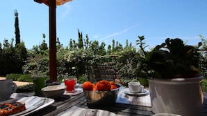 Ein Tisch draußen mit Pizza, Früchten und einem Getränk, unter einer hölzernen Pergola mit blauem Himmel im Hintergrund.