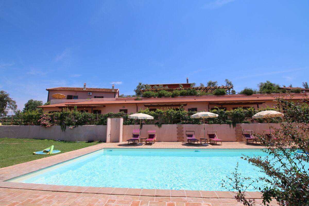 A swimming pool with lounge chairs and umbrellas, surrounded by a landscaped garden and a large building with a red roof.