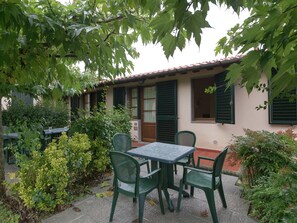 Un espace patio aménagé avec une table et des chaises, entouré de verdure et bordé d’un bâtiment doté de volets.