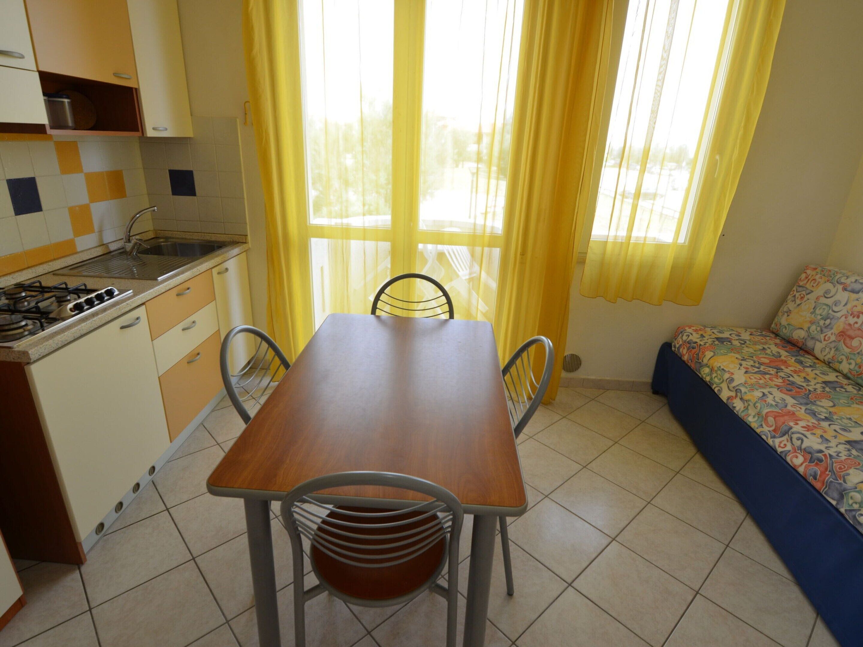 A compact kitchen with a stove, sink, and yellow curtains. A dining table with chairs is in the center. A sofa with a colorful blanket is against the wall.