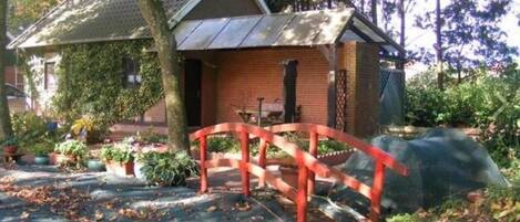 Una pequeña casa de ladrillo con techo metálico, un puente rojo y un jardín con diversas plantas.