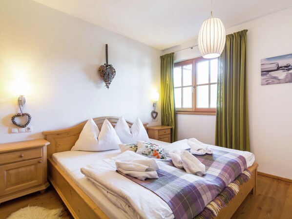 A neatly made bed with white linens and a plaid blanket, wooden headboard, bedside tables with lamps, a wall-mounted light fixture, and a window with green curtains.