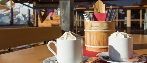 Dos tazas blancas de café batido sobre una mesa de madera con una cuchara y un pequeño paquete, frente a un interior de madera y una ventana con vista a un edificio.