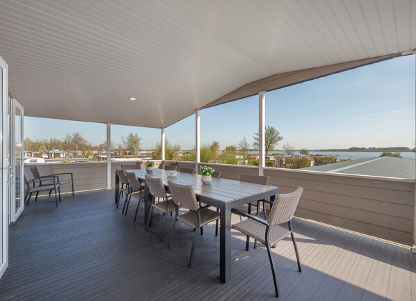A covered outdoor patio with a dining table and chairs, large windows offering a view of a body of water and surrounding landscape. - Group Retreat in Giethoorn (Giethoorn)