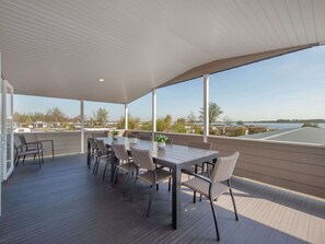 A covered outdoor patio with a dining table and chairs, large windows offering a view of a lake and buildings.