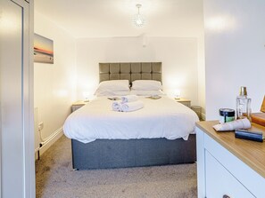 Un lit bien fait, avec du linge de lit blanc et des serviettes pliées, une tête de lit rembourrée de couleur grise, des tables de chevet avec des lampes et une petite table d’appoint en bois sur laquelle sont posés des articles de toilette.