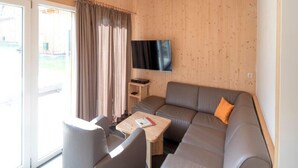 A modern living room with a grey sofa, a small table, a TV mounted on the wall, and a sliding glass door leading to an outdoor area. - Chalet in the ski Area in Styria (Sankt Georgen am Kreischberg)