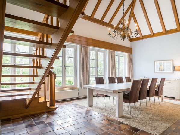 A spacious dining area with a high ceiling and exposed wooden beams, large windows with curtains, and a chandelier. - Peaceful Farmhouse in De Cocksdorp near Sea (De Cocksdorp)