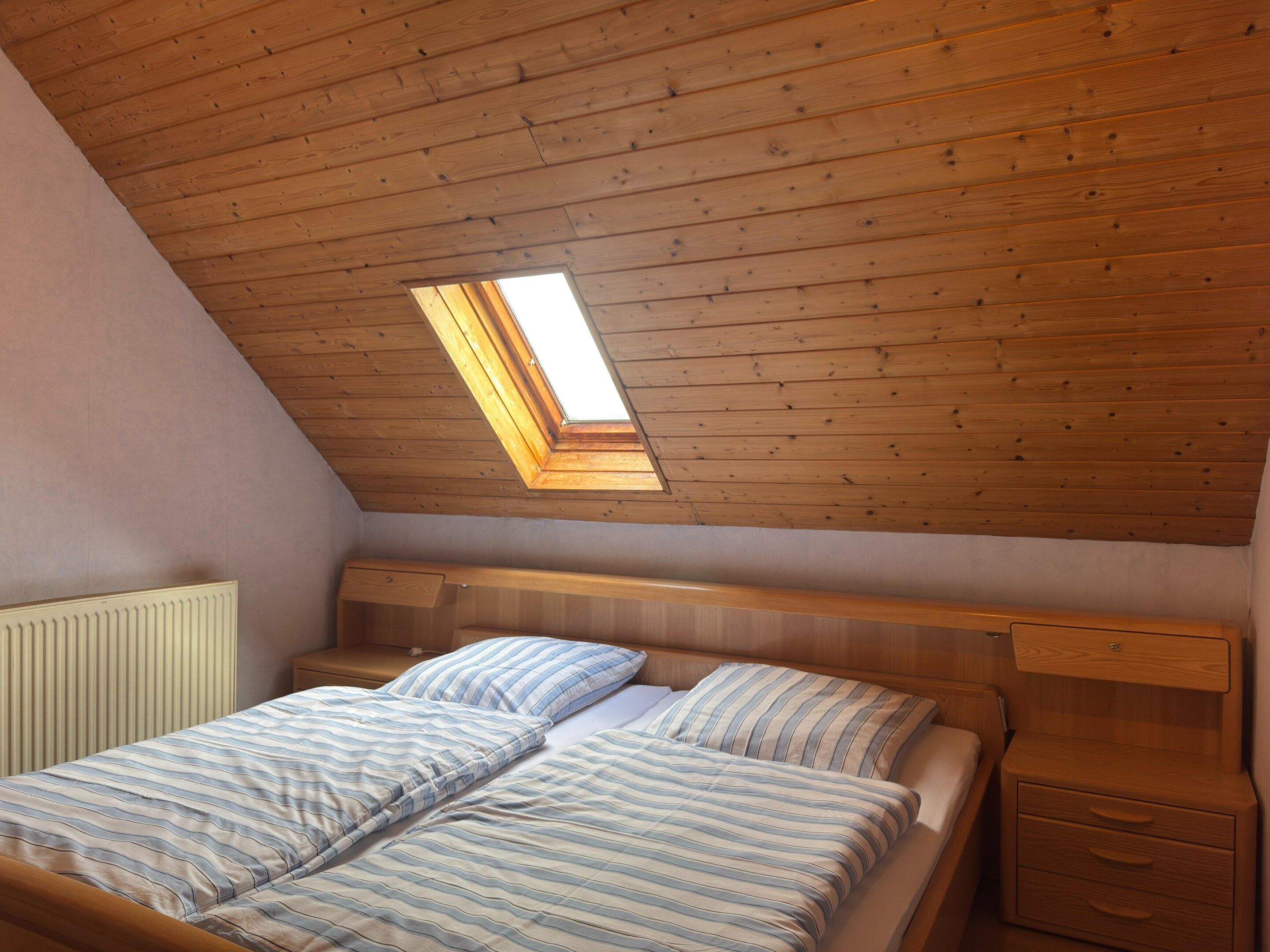 Un dormitorio con techo inclinado, tragaluces, cama con ropa de cama a rayas y una mesita de noche con cajones.