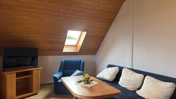A room with a sloped ceiling, a television on a wooden stand, a blue armchair, a wooden coffee table with a flower arrangement, and a dark blue sofa with white cushions.