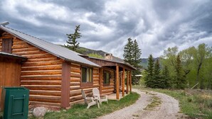 Una baita di legno con un portico, un vialetto sterrato che vi conduce e una foresta circostante.