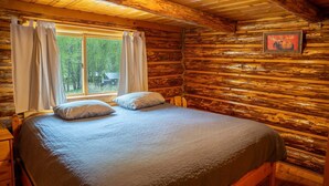 Camera da letto in una baita di legno, con un letto, due cuscini, un comodino, una cassettiera, una finestra con tende e un quadro appeso al muro.