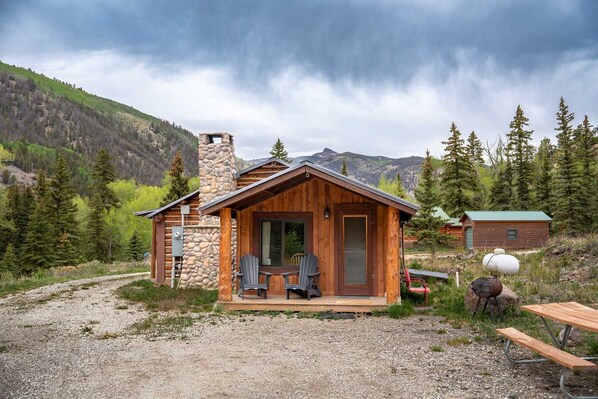 A rustic cabin with a stone chimney, wooden exterior, and a porch with two Adirondack chairs. - Keepin' It Reel - Large Cabin for the Whole Family at Historic Vicker's Ranch - Sleeps 10 (Lake City)