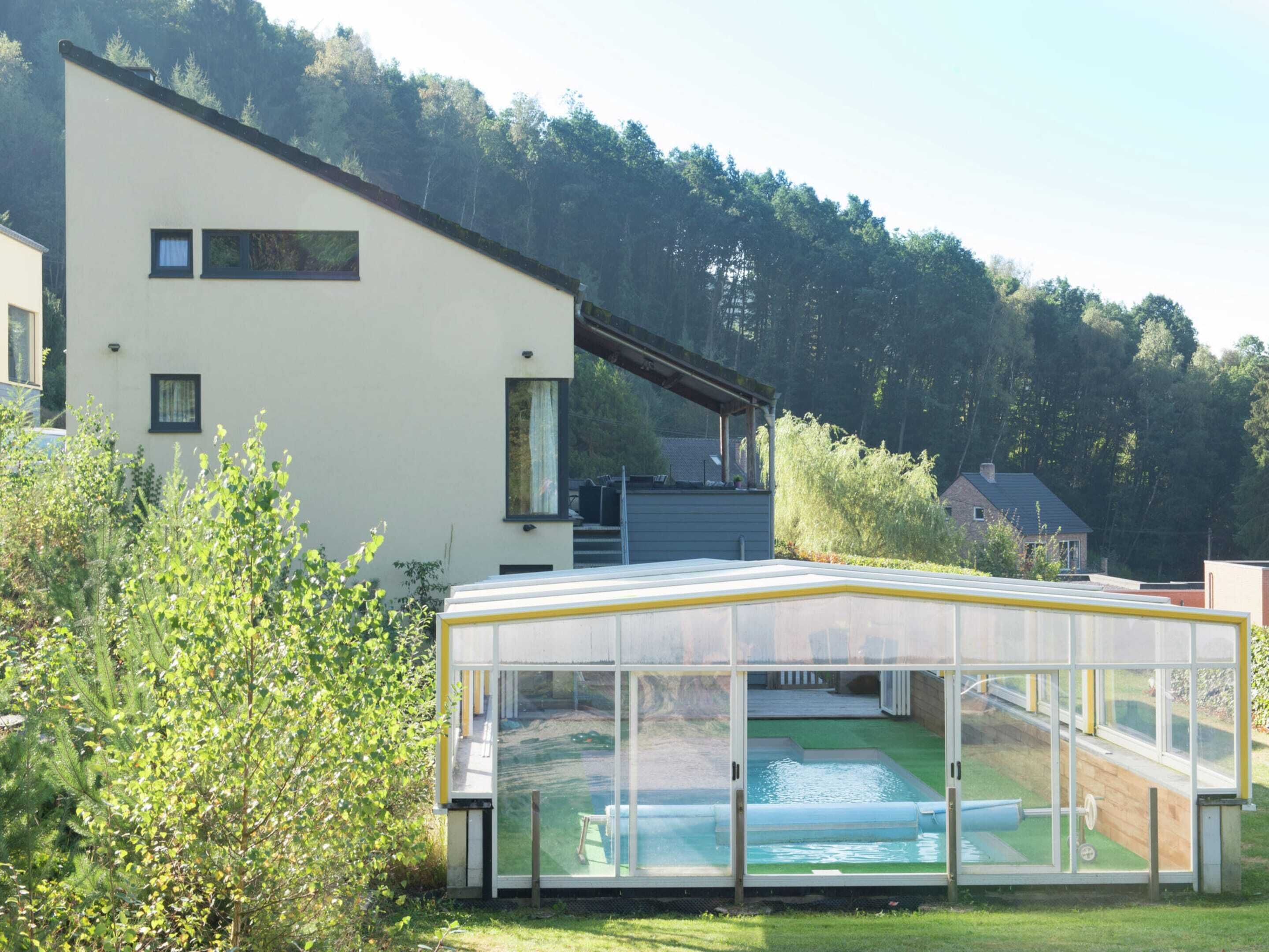 Une maison moderne dotée d’un espace piscine entièrement vitré, entourée de verdure et bordée par une colline boisée.