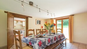 A dining area with a table set for four, wooden chairs, a bench, a vase with flowers, and a kitchen visible through an open door. - Rustic Cottage in Jevigne (Lierneux)