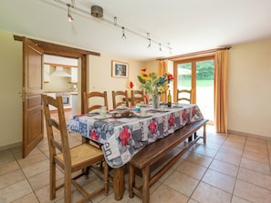 A dining area with a table set for four, wooden chairs, a bench, a vase with flowers, and a kitchen visible through an open door. - Rustic Cottage in Jevigne (Lierneux)