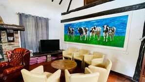 A living room with a TV, leather armchair, beige sofas, and a painting of cows on the wall. - Farmhouse in Sint Joost With Pool & BBQ (Sint Joost)