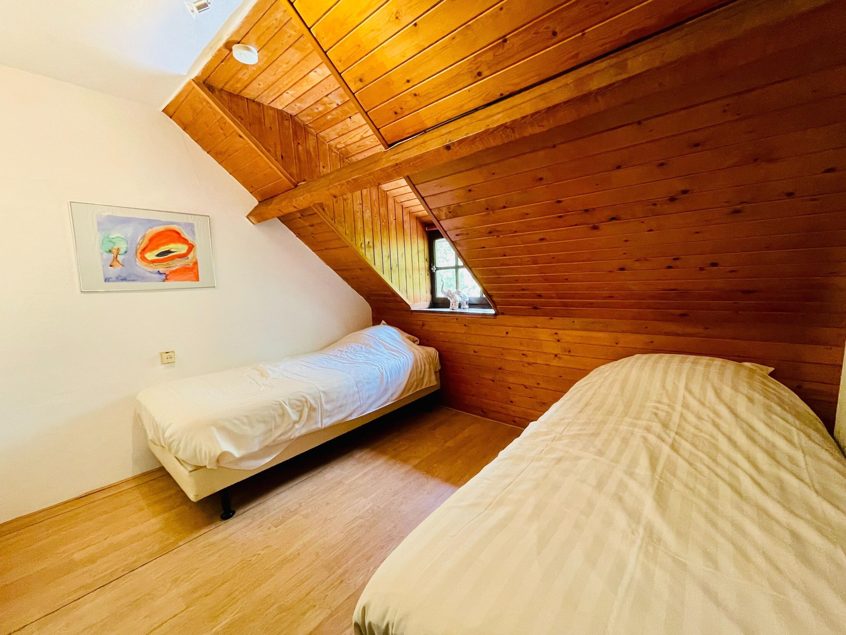 A room with wooden sloped ceilings, two beds, a window, and a painting on the wall.