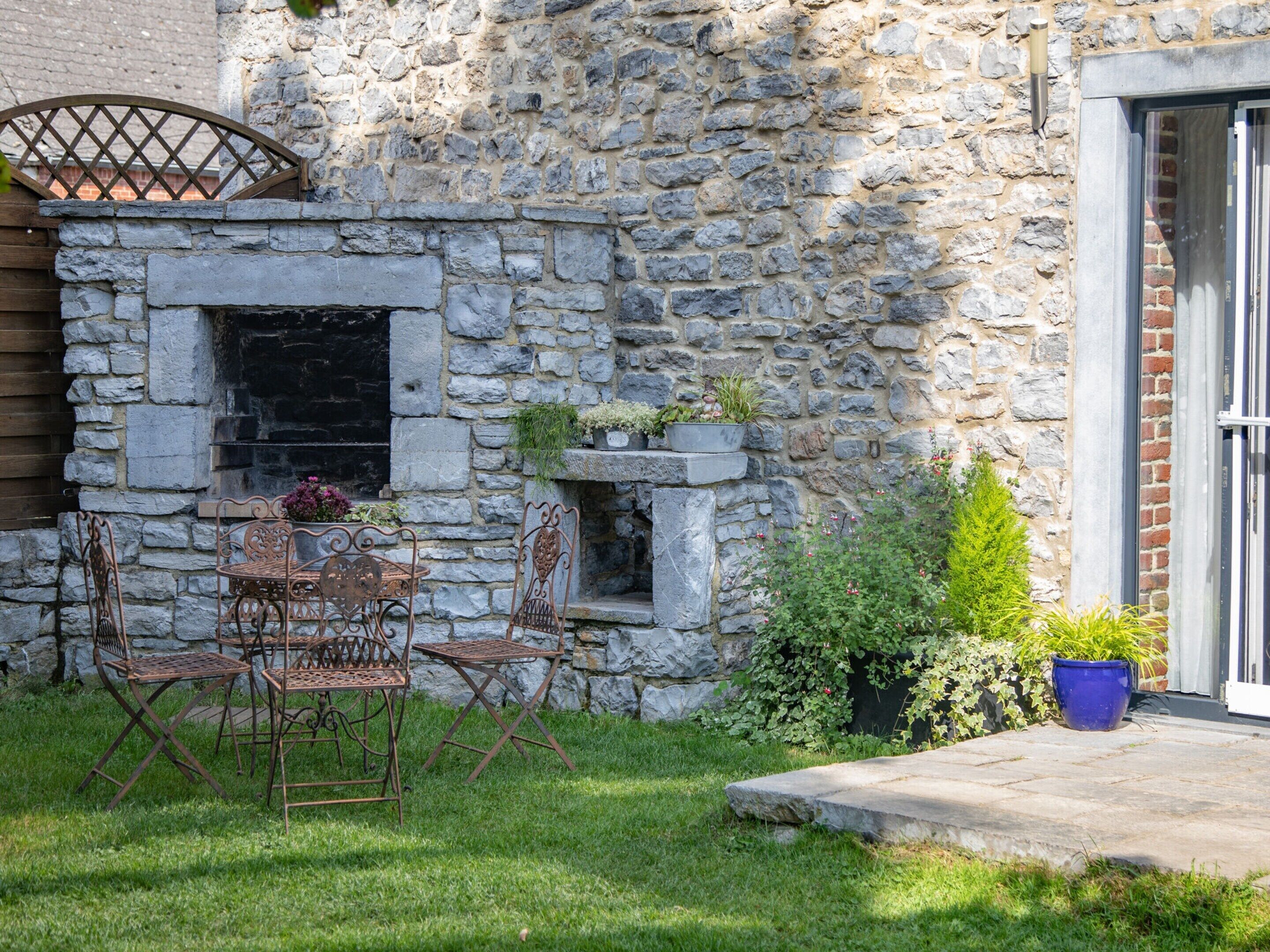 Un mur en pierre avec un barbecue intégré, un espace de détente extérieur avec des chaises en métal et un pot de fleurs.