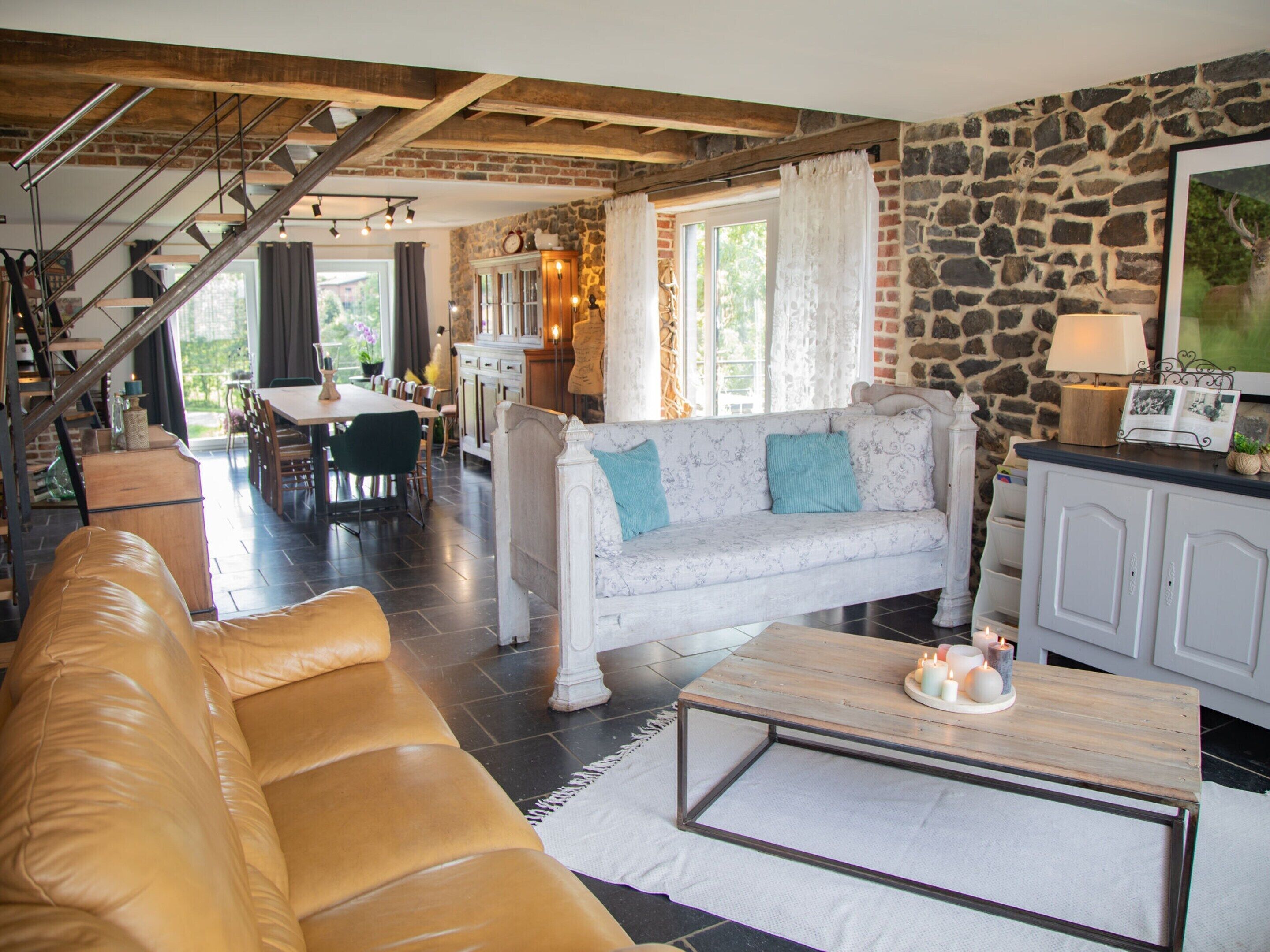 A spacious living area with a leather sofa, a white patterned sofa, and a coffee table with candles. A staircase with exposed beams leads to a dining area.