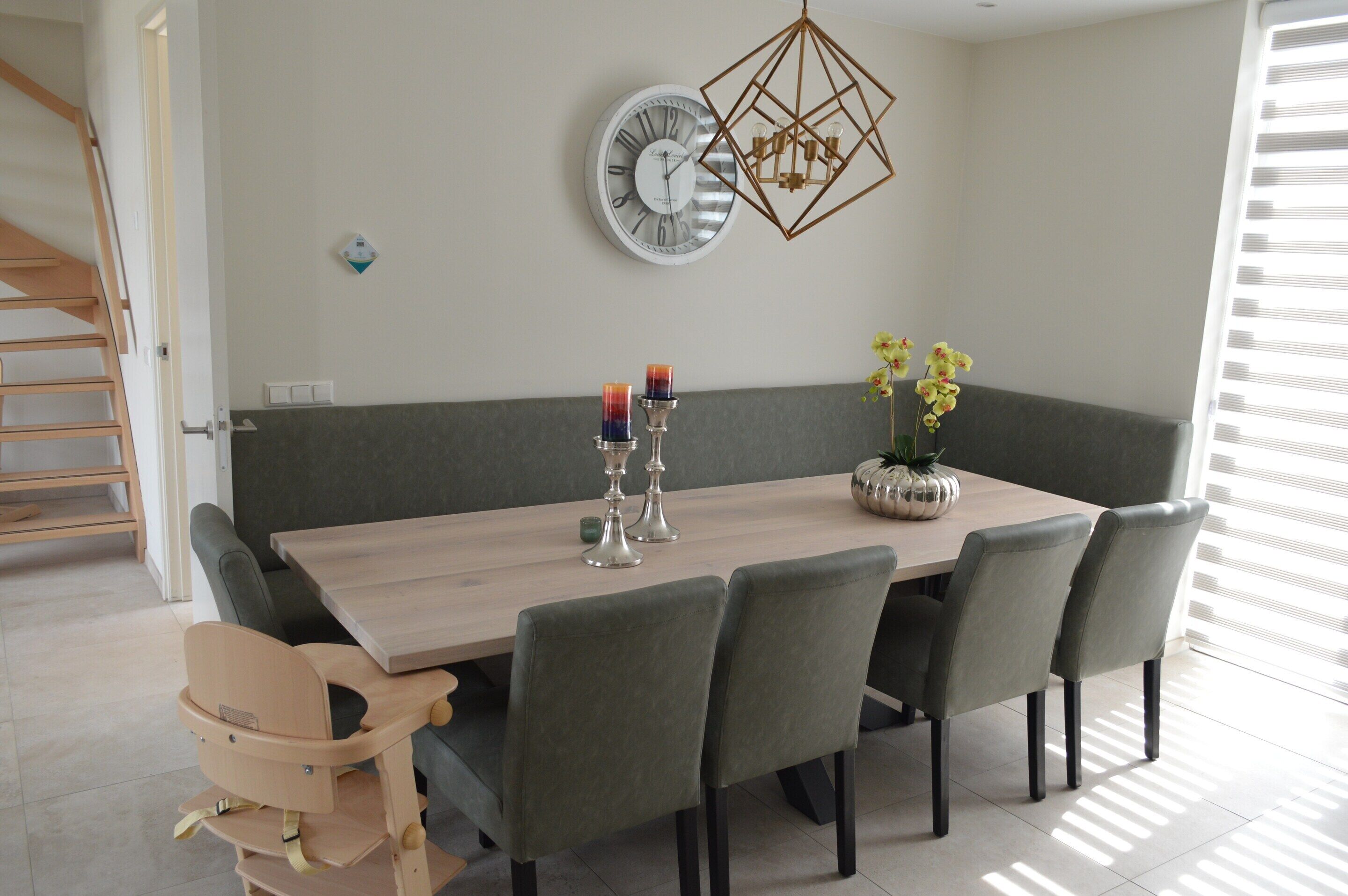 Een eetruimte met een houten tafel, zes stoelen, een bank, een wandklok, een geometrische lamp en een vaas met bloemen.