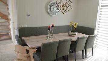 Een eetruimte met een houten tafel, zes stoelen, een bank, een wandklok, een geometrische lamp en een vaas met bloemen.