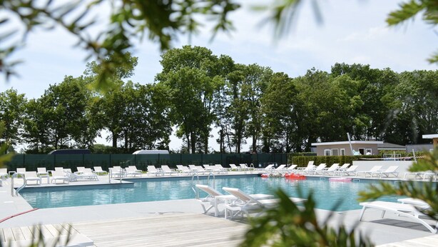 Una piscina con sillones blancos, rodeada de árboles y un edificio al fondo.