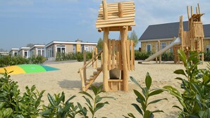 Un parque infantil con estructuras de madera, incluyendo un tobogán y elementos para escalar, situado sobre un suelo arenoso y rodeado de vegetación.