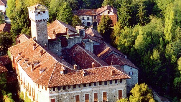 Vista aérea de un edificio histórico con una torre, múltiples tejados y un patio rodeado de árboles.