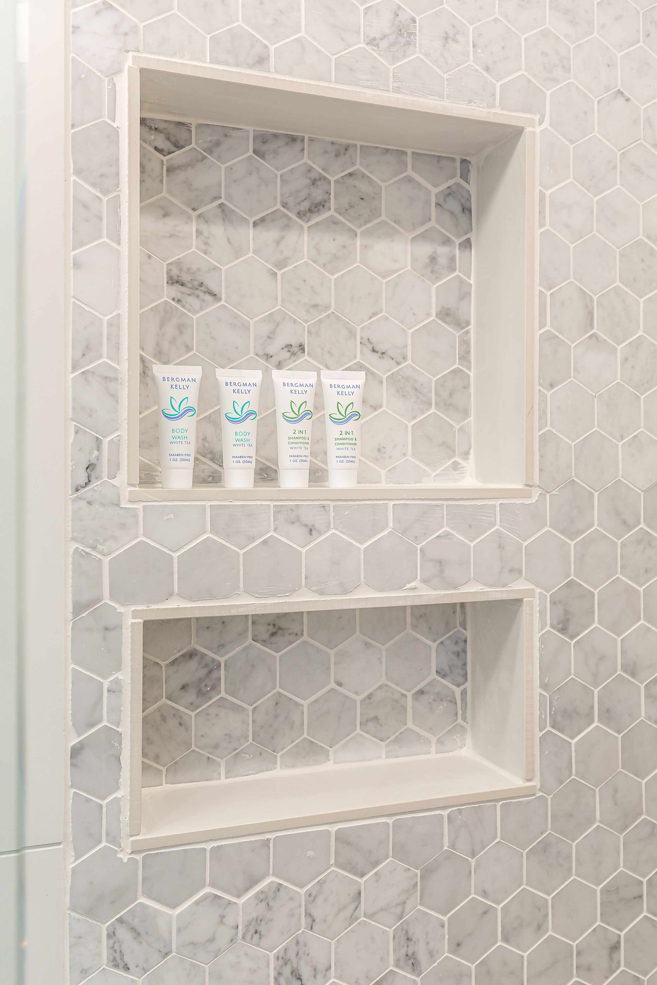 Un baño con baldosas de mármol hexagonales y dos estantes blancos con botellas de Bergman Kelly de jabón líquido y champú.