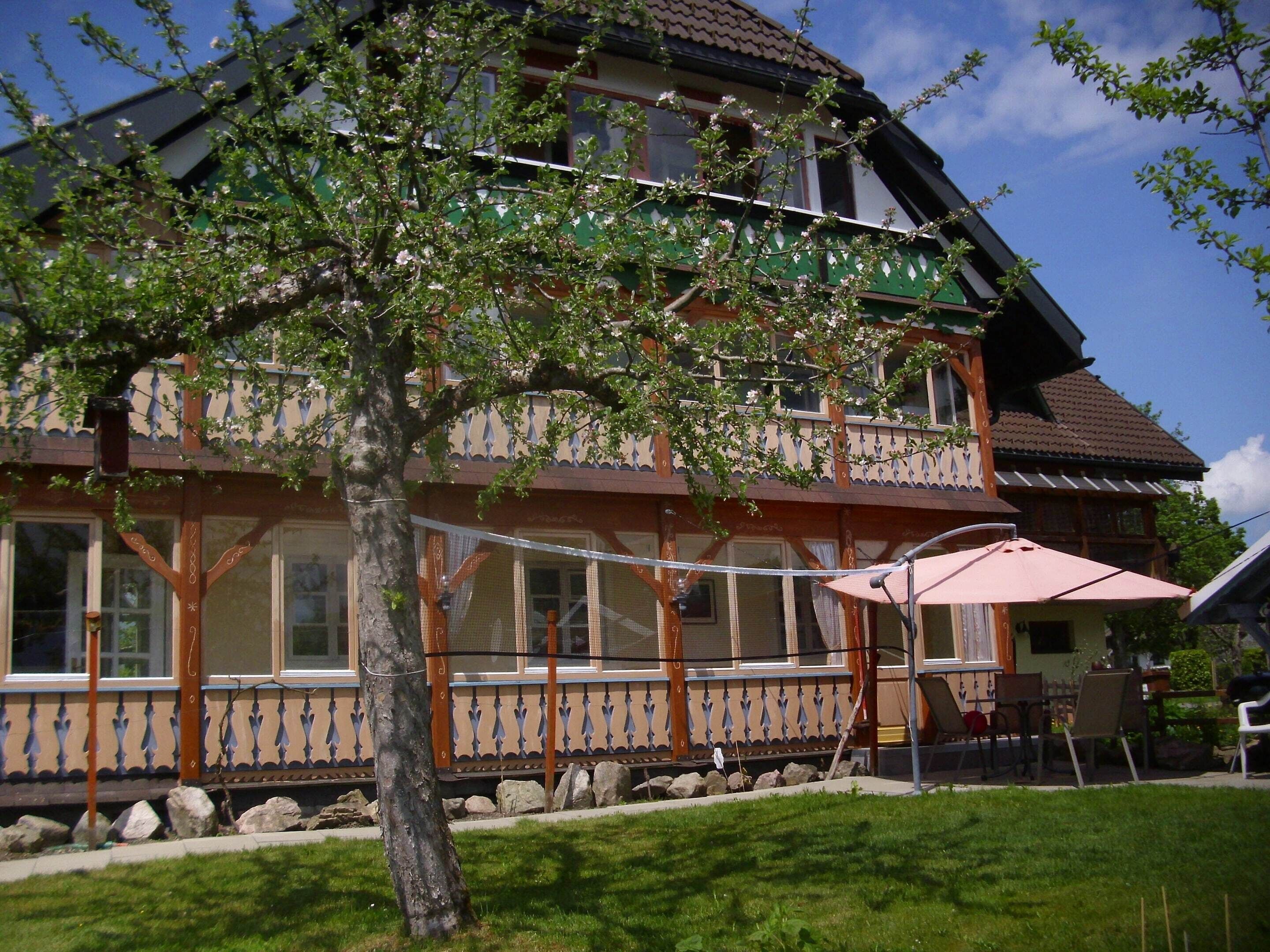 A traditional chalet-style building with a balcony, a tree in the foreground, and a clear sky.