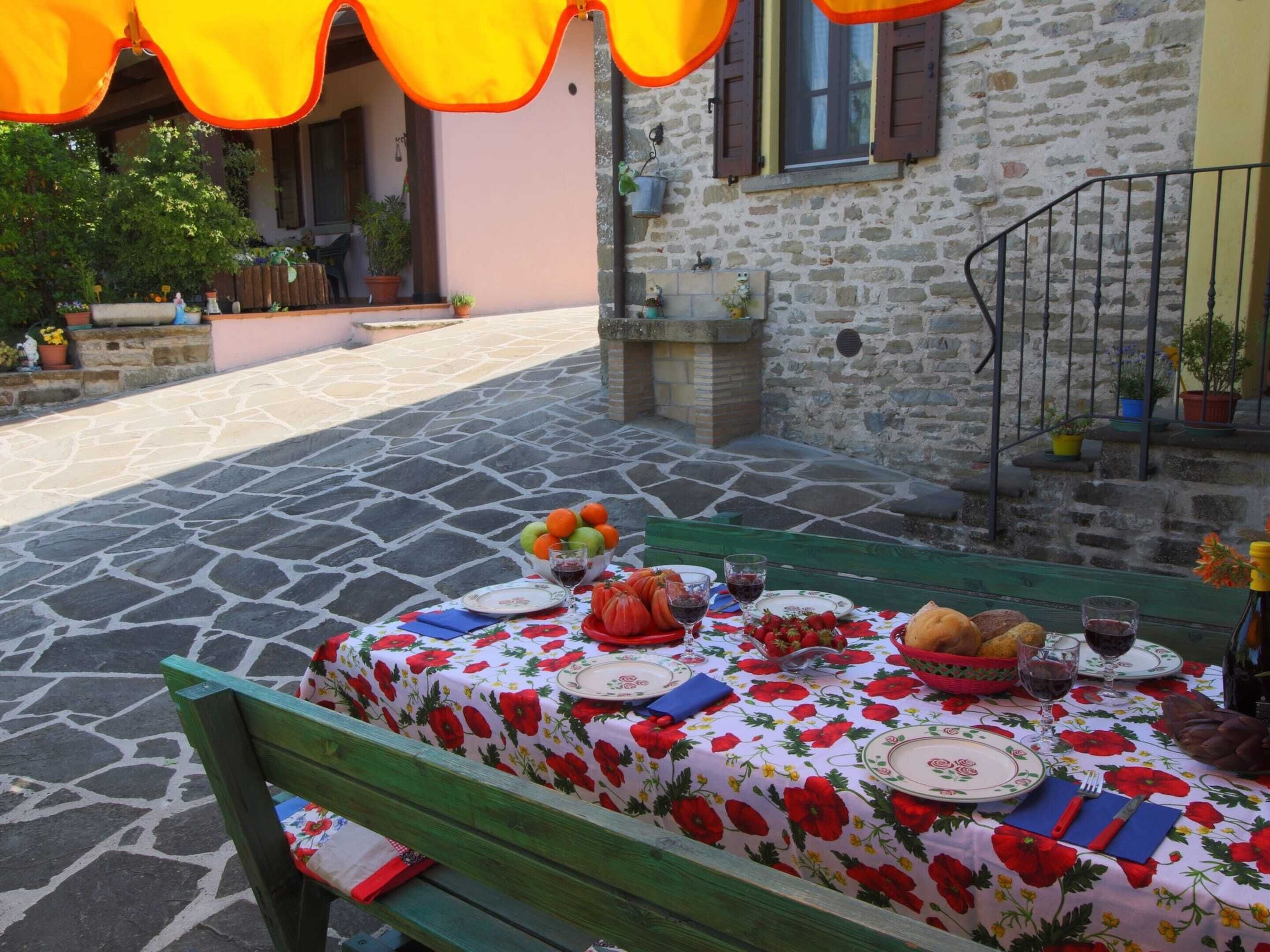 A table set outdoors with a floral tablecloth, plates, cutlery, and a bowl of fruit.