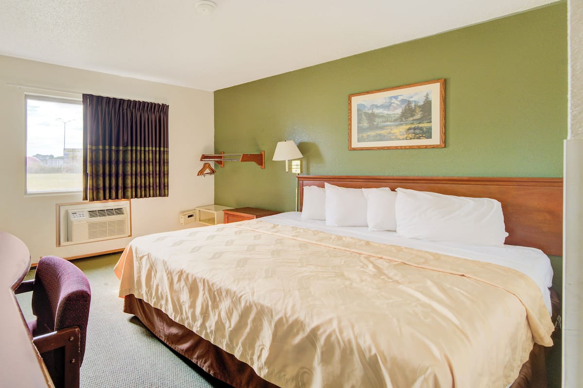 A hotel room with a large bed, a chair, a desk, an air conditioning unit, a lamp, and a painting on the wall.
