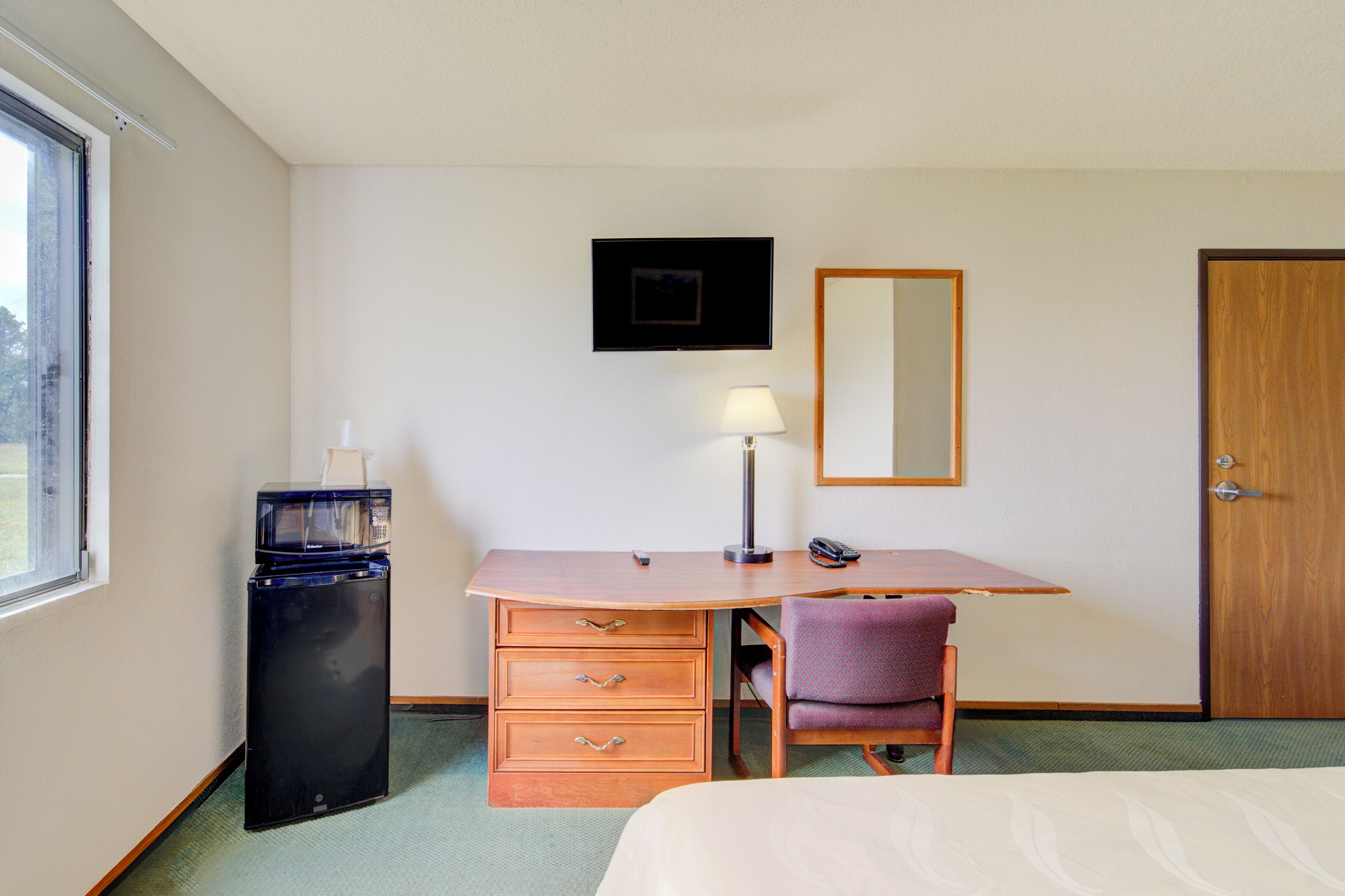 A hotel room with a bed, a desk with a chair, a small refrigerator, a lamp, a mirror, and a door.