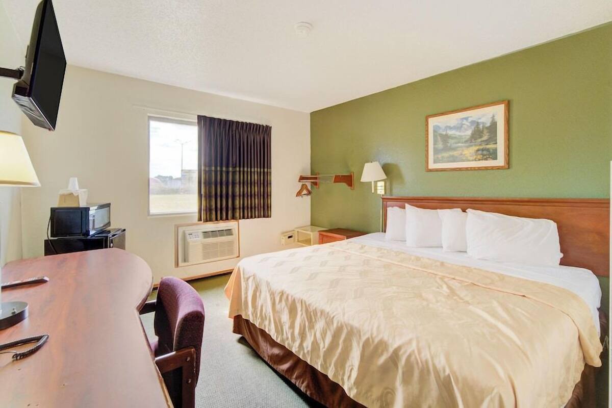 A hotel room with a bed, a desk, a television, a window with curtains, an air conditioner, and a painting on the wall.