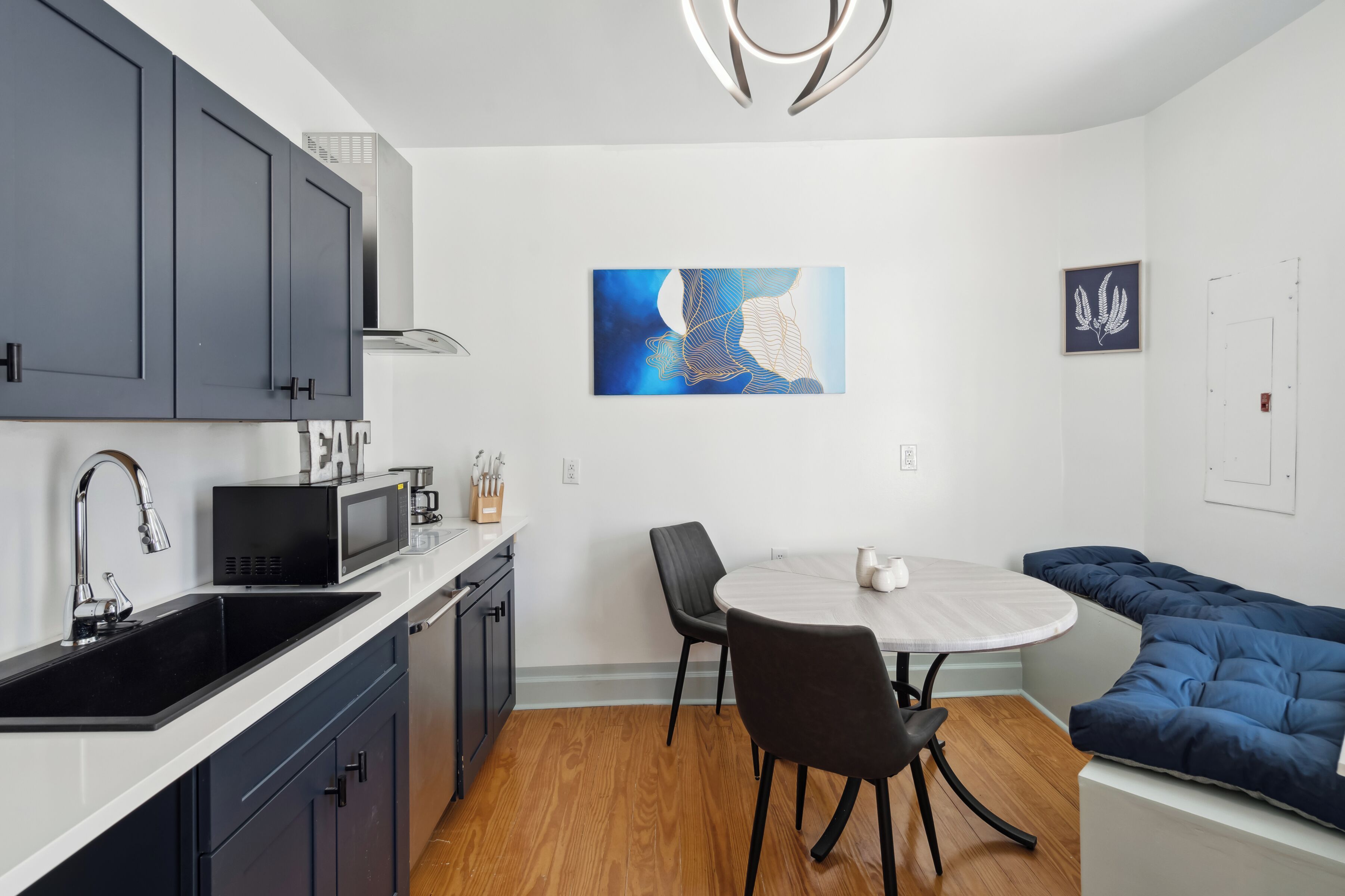 A modern kitchen with dark cabinets, a microwave, and a sink. There is a round dining table with chairs and a blue sofa. A painting is on the wall.