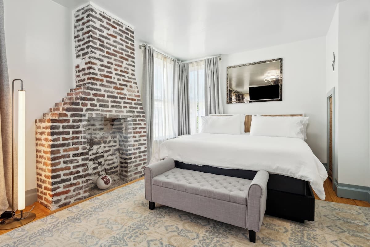 A bedroom with a brick fireplace, a bed with white linens, a grey bench, and a patterned rug.