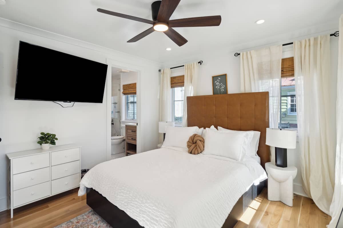 A bedroom with a large bed, a ceiling fan, a TV mounted on the wall, a bedside table with a lamp, and a window with curtains.