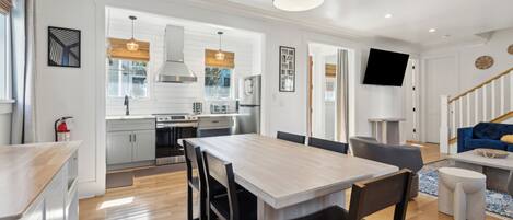 A modern kitchen with white cabinets, a dining area with a wooden table and chairs, a living room with a blue sofa, and a staircase with wooden railings.