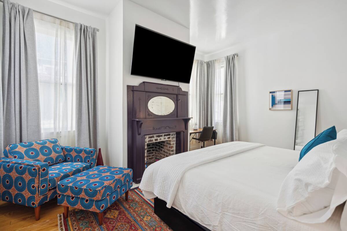 A bedroom with a bed, a television mounted above it, a fireplace, and a seating area with a patterned chair and ottoman.