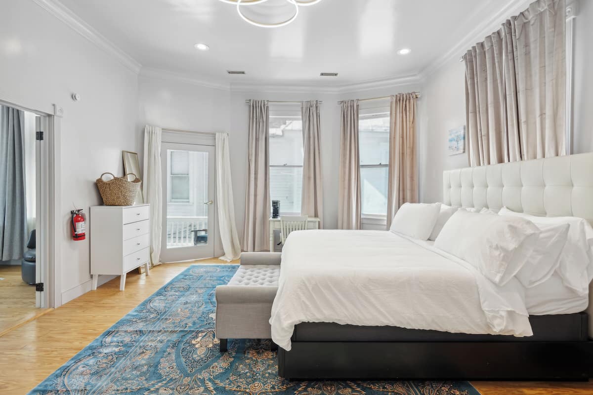 A bedroom with a large bed, a white dresser, a patterned rug, and a view of the outdoors.