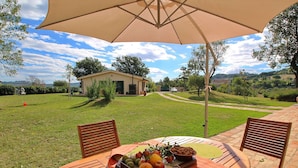 An outdoor dining area with a table set for two, a large umbrella, and a view of a house and landscape in the background. - Cottage in Montelabbate With Pool & Forest (Montelabbate)