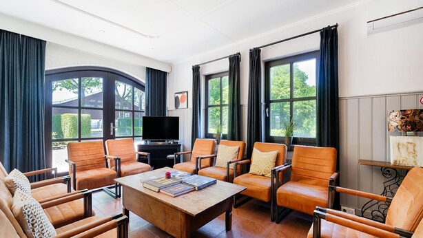 Ein geräumiges Wohnzimmer mit Ledersesseln, einem Holztisch und einem Flachbildfernseher. Große Fenster bieten Blick ins Grüne.
